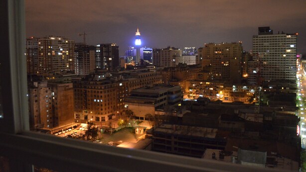 A nighttime cityscape with high-rise buildings, a prominent illuminated tower, and a view through a window.