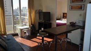 A compact living space with a sofa, a small dining table with stools, a television, and a city view through large windows.