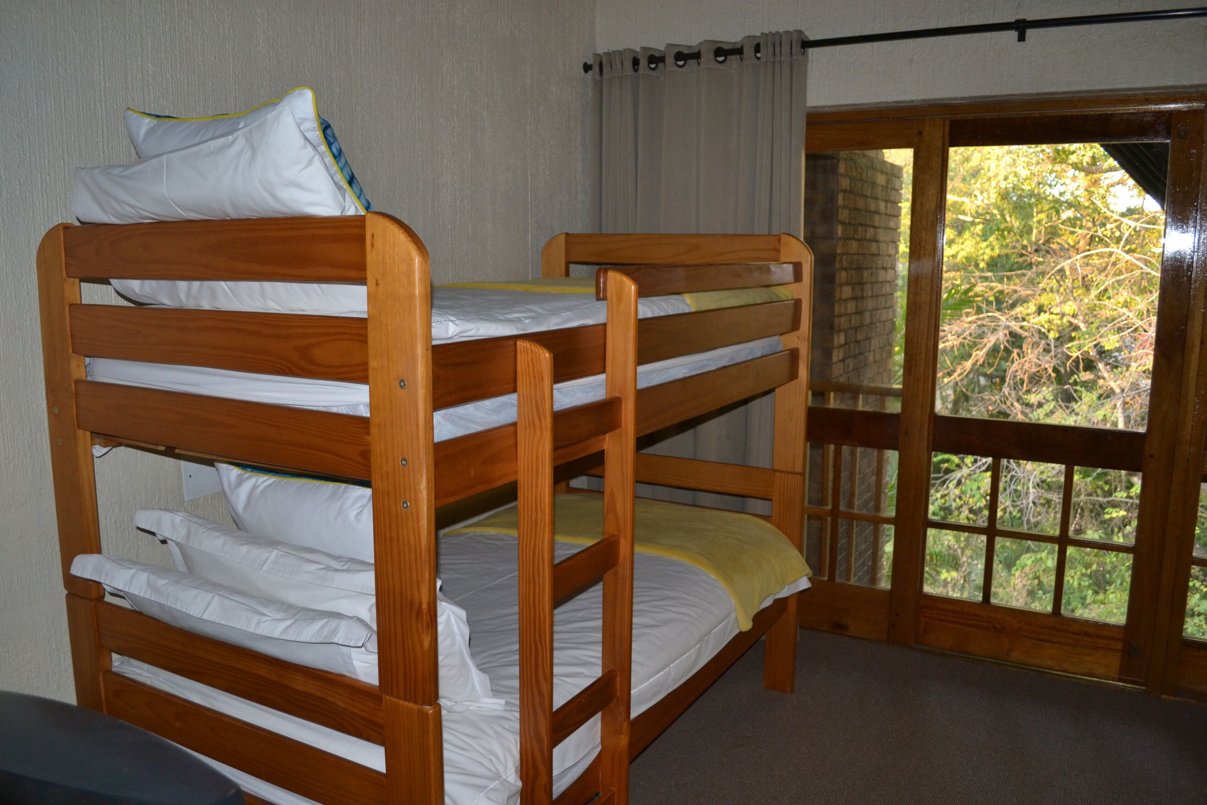 A bunk bed with white bedding and a yellow blanket, a window with curtains, and a view of greenery outside.