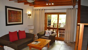A living room with a brown sofa, a wooden coffee table, and a brown armchair. There is a painting on the wall and a window with curtains. - Kruger Park Lodge - Golf Safari SA (Mbombela)