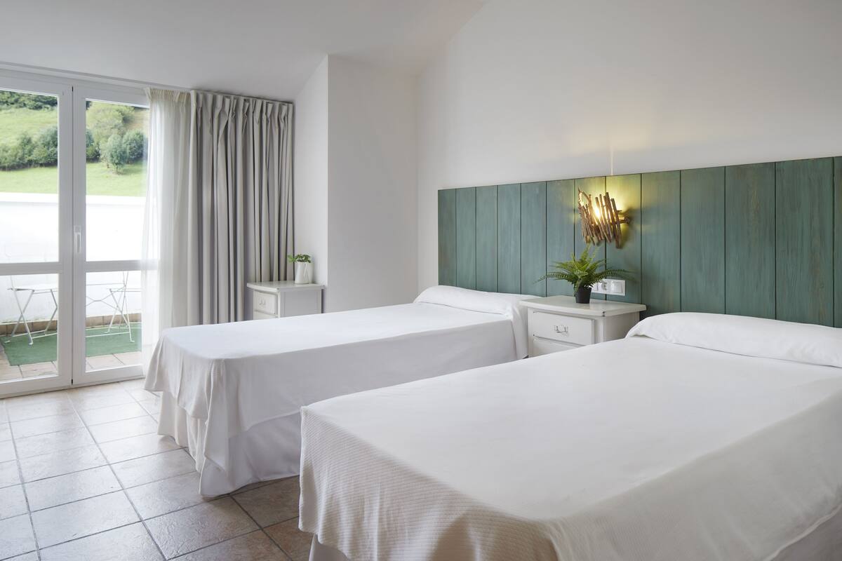 A hotel room with two beds, a sliding glass door to the outside, a small table with a plant, and a wall-mounted light fixture.