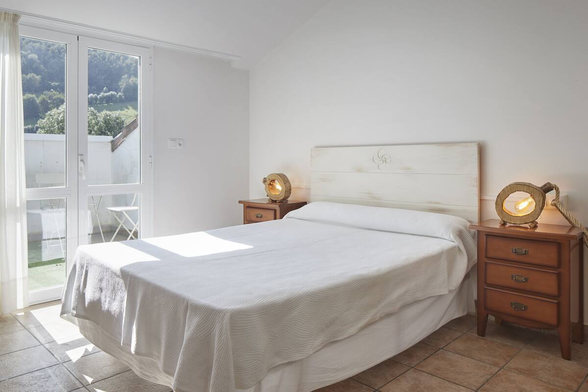 A bedroom with a large bed, a wooden headboard, two bedside tables with lamps, and a sliding glass door leading to a balcony.