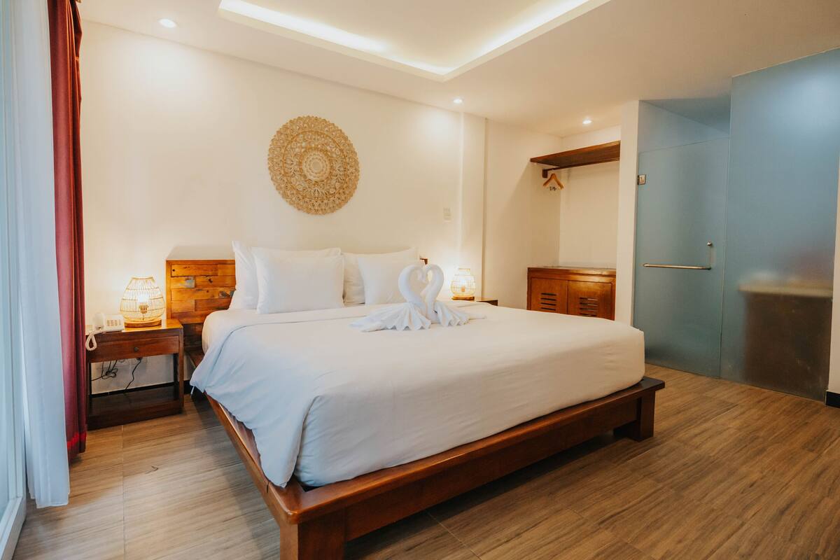 Una cama bien hecha con sábanas blancas, un cabecero de madera y un adorno decorativo en la pared. Hay una mesita de noche con una lámpara y un teléfono. El piso es de madera y la puerta es azul.