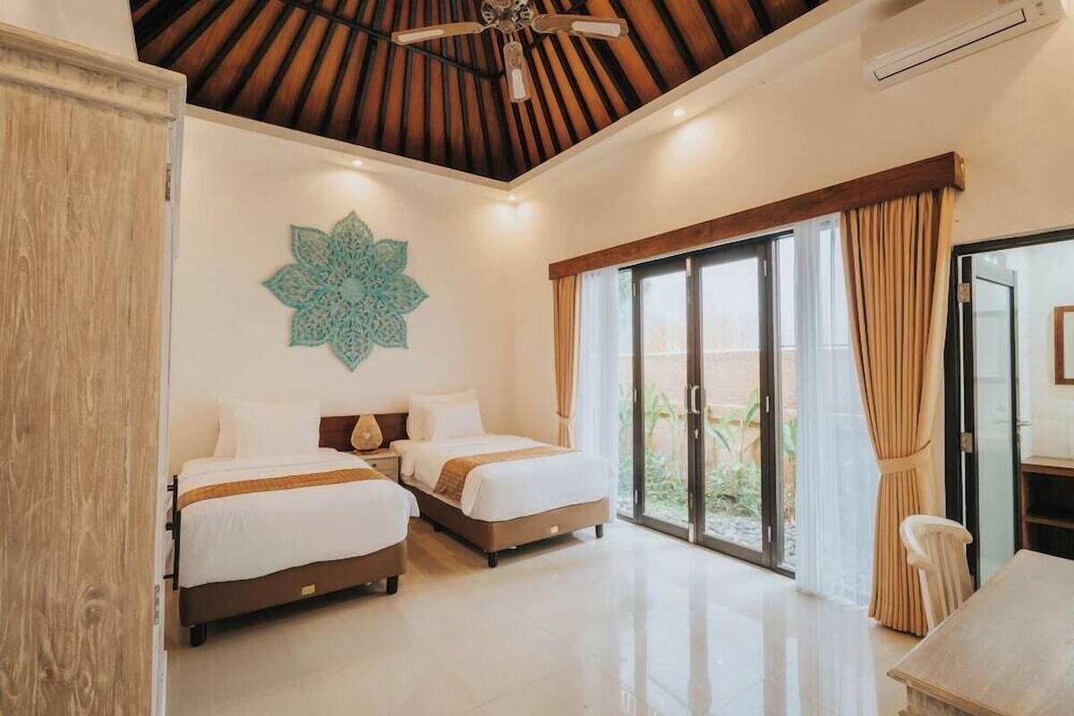 Habitación de hotel con dos camas, un ventilador de techo, un ventanal grande con cortinas y un adorno en la pared.