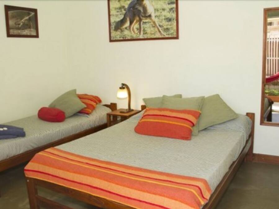 Quarto com duas camas de solteiro, cada uma com travesseiros e cobertor. Há uma mesa de cabeceira com luminária entre as camas.