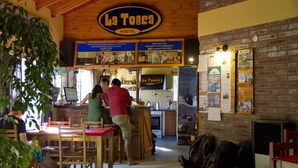 A hostel interior with a wooden ceiling, brick walls, and a bar area. There are wooden tables and chairs, and a person is seen at the bar. - La Tosca Hostel (Puerto Madryn)