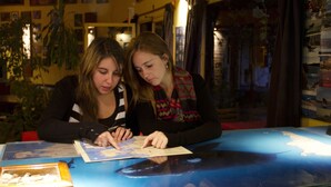 Two individuals examining a map at a table with a glass top, surrounded by various framed pictures and a plant. - La Tosca Hostel (Puerto Madryn)