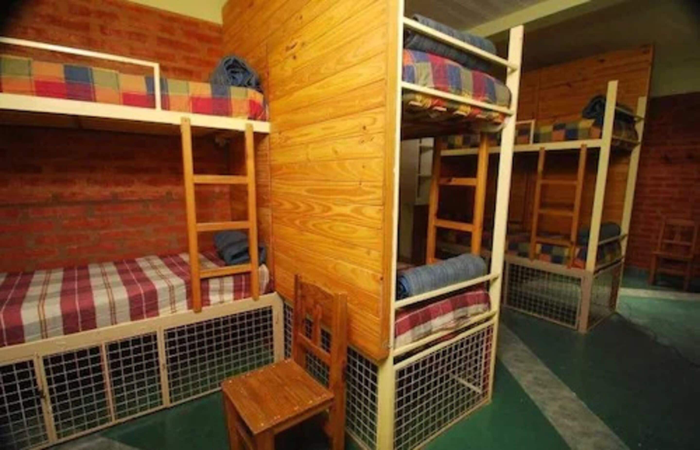 Dormitorio con literas de madera, barandillas metálicas y una pequeña silla de madera.