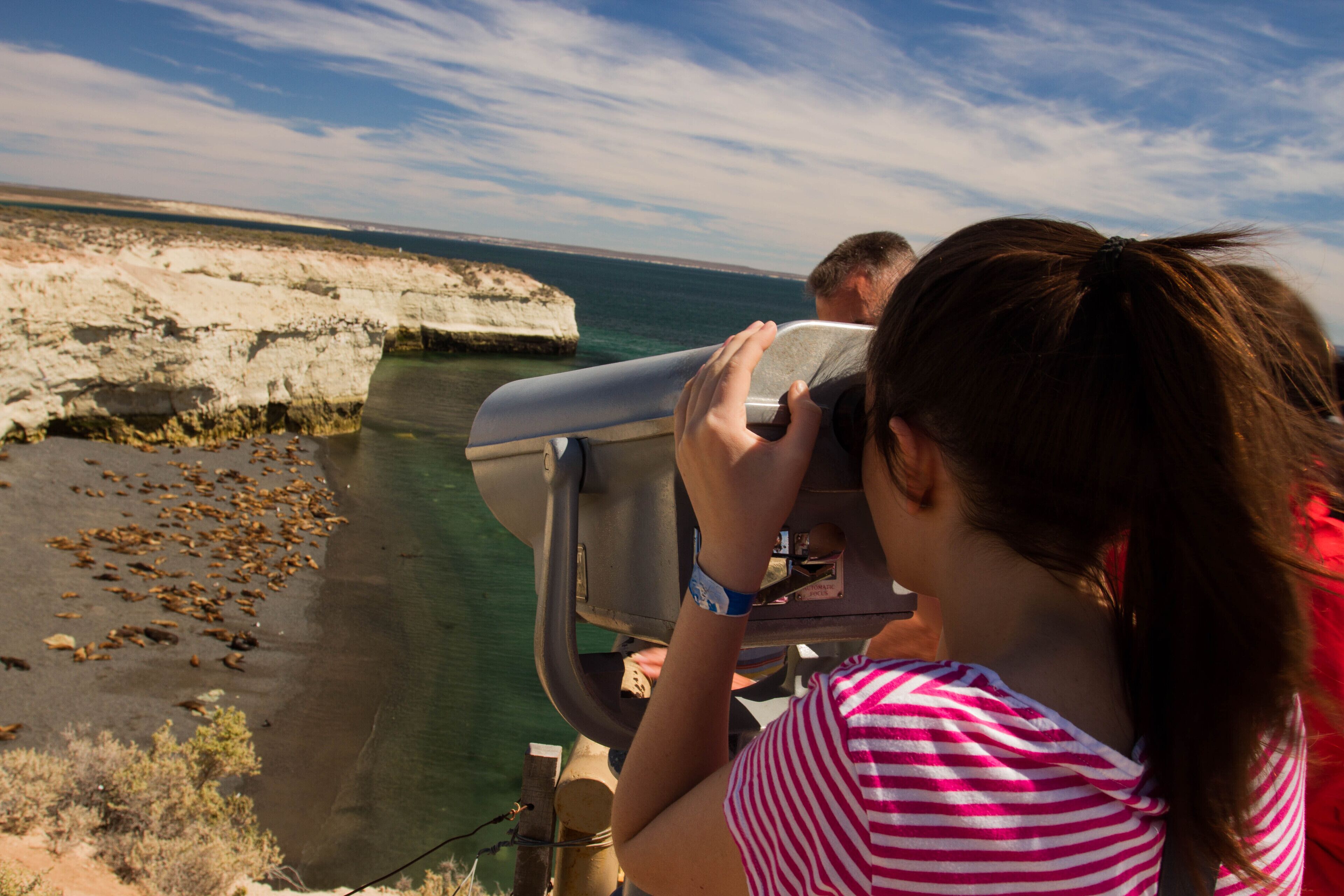 A person using binoculars overlooking a coastal landscape with cliffs and clear water.