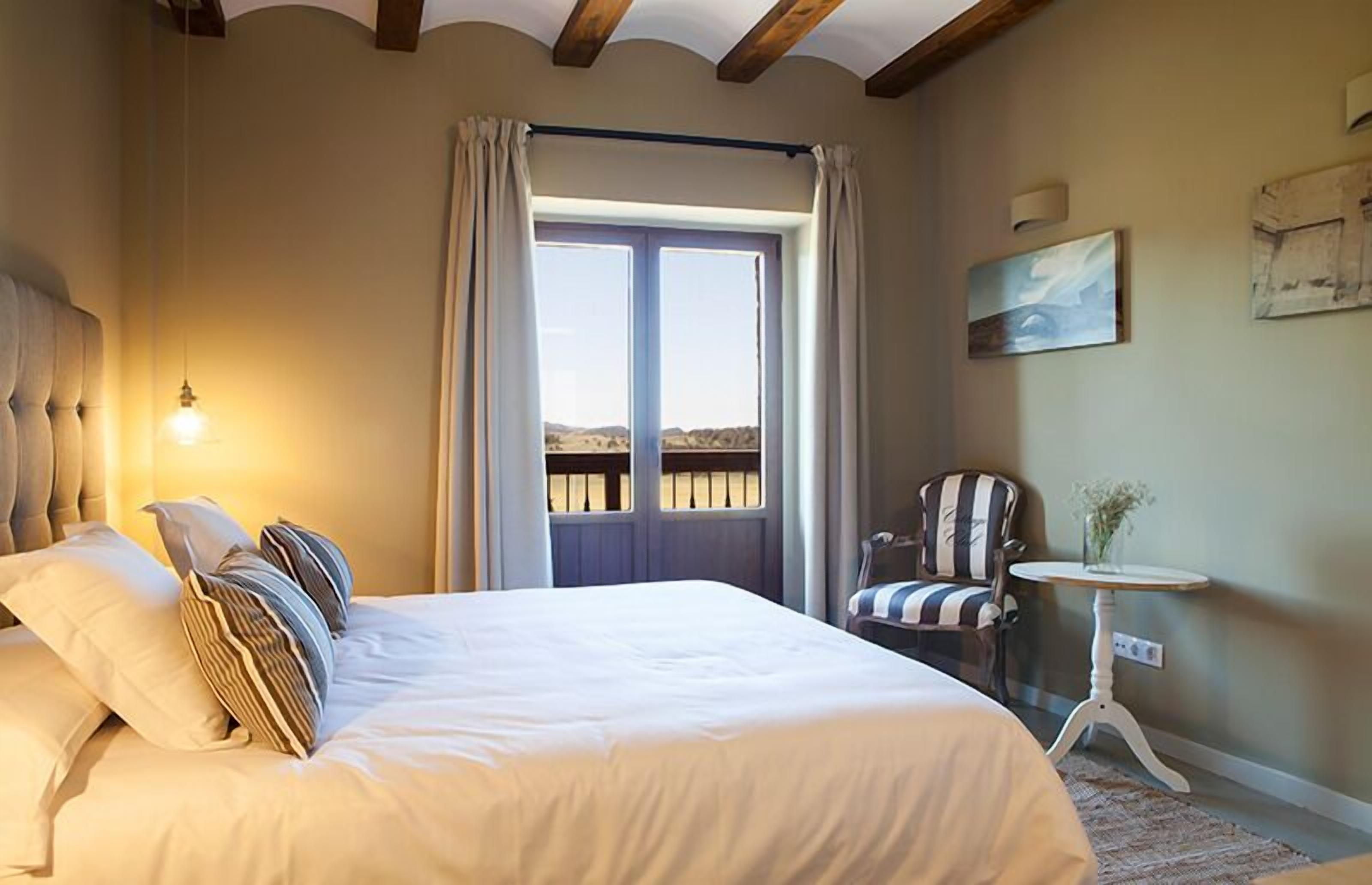 A bedroom with a large bed, a chair, a small table, and a window with curtains.
