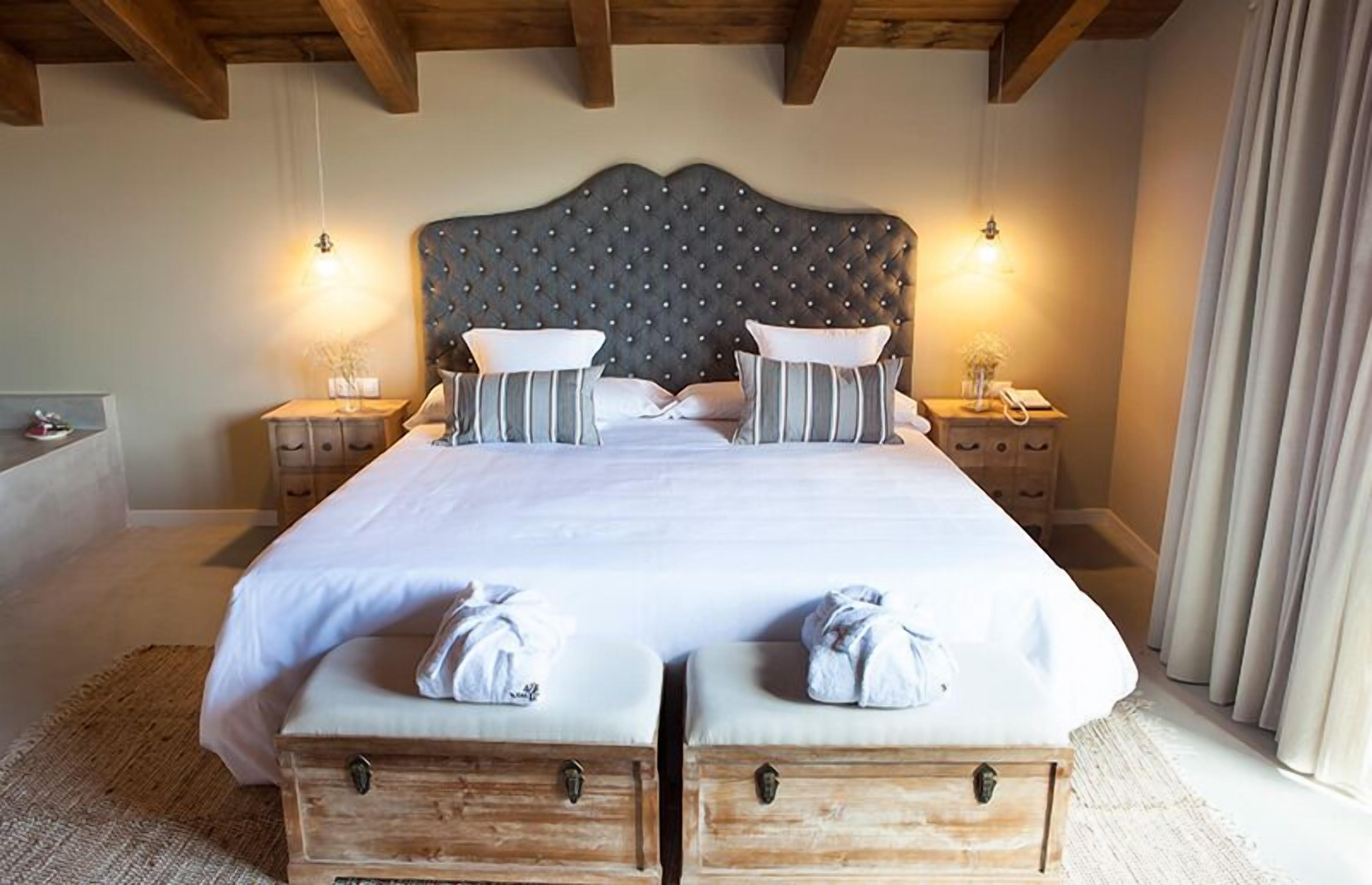 A double bed with a tufted headboard, two bedside tables with lamps, and a large window with curtains.
