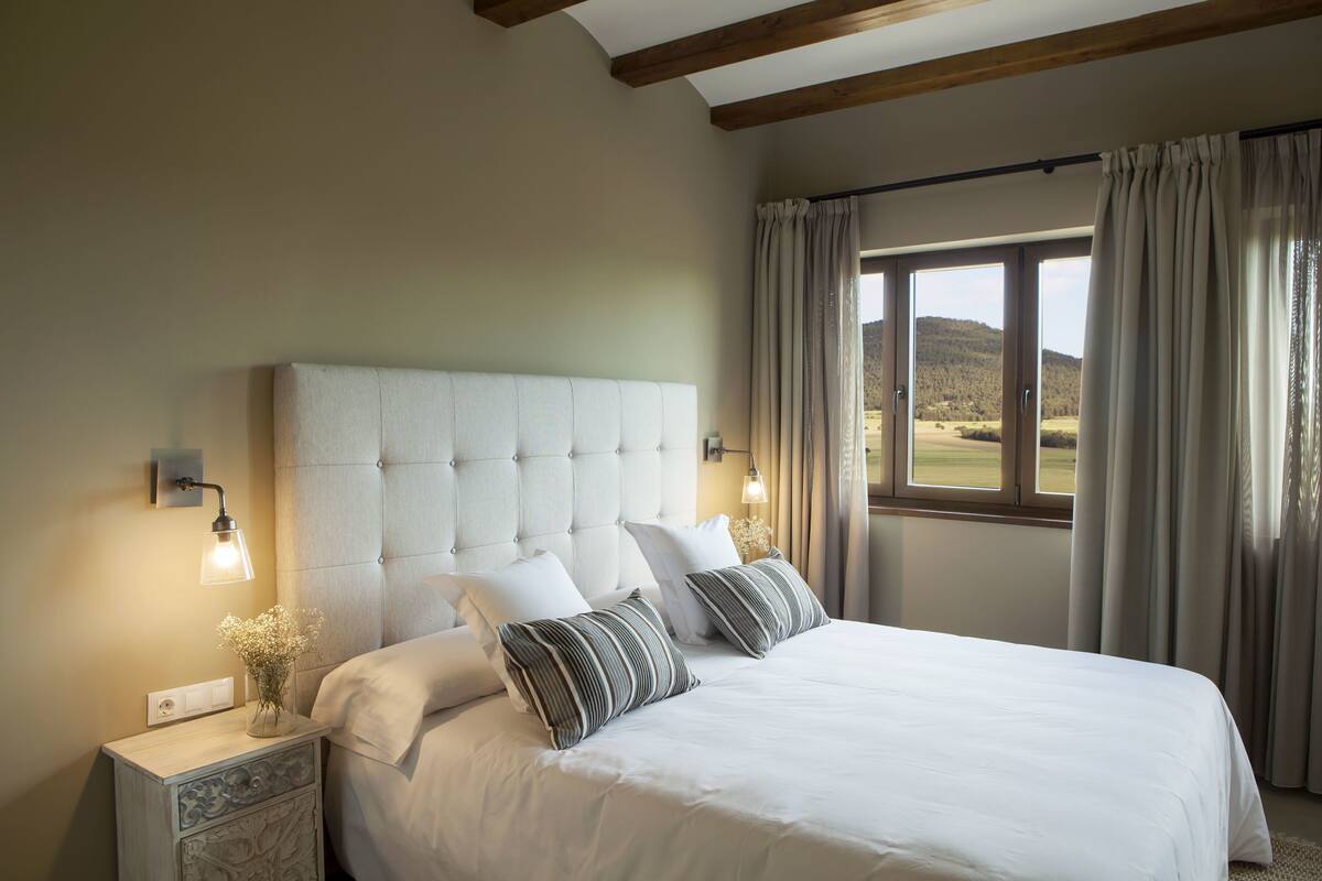 A bedroom with a large bed, two bedside tables, a nightstand, a window with curtains, and a view of the outdoors.
