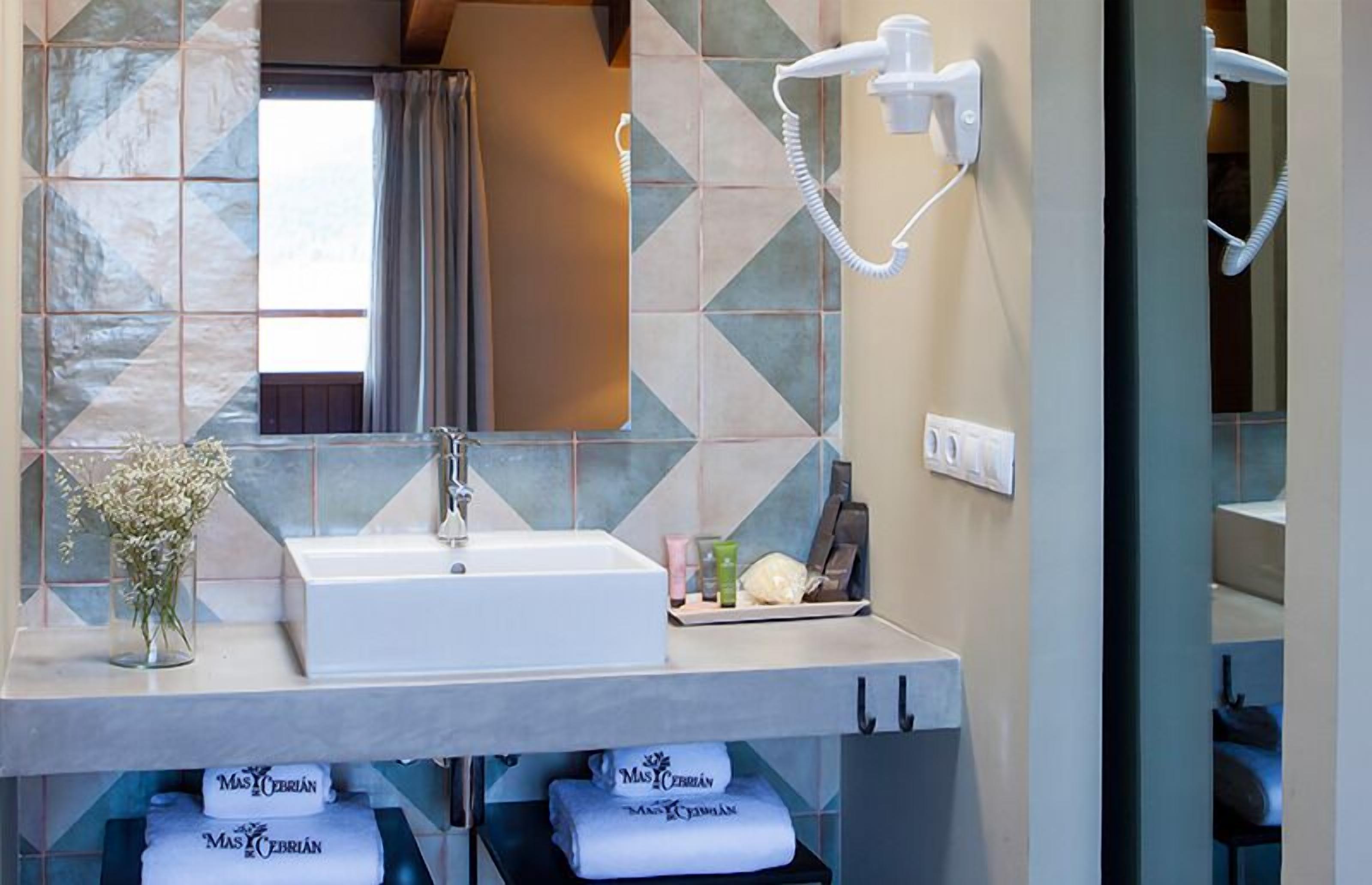 Un bany amb un mirall gran, un lavabo, un assecador d'eixugat i tovalloles amb la marca "Mas Cerrian".
