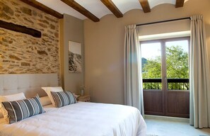 A bedroom with a stone wall, a bed with striped pillows, a nightstand, and a view of greenery through a sliding door. - Hotel Mas de Cebrián (Puertomingalvo)