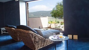 A room with a wicker chair, a small round table with drinks, and a view of a mountainous landscape through large windows. - Hotel Mas de Cebrián (Puertomingalvo)