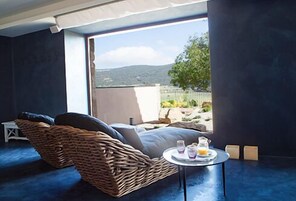 Una habitación con una silla de mimbre, una mesita redonda con bebidas y vistas a un paisaje montañoso a través de amplias ventanas.
