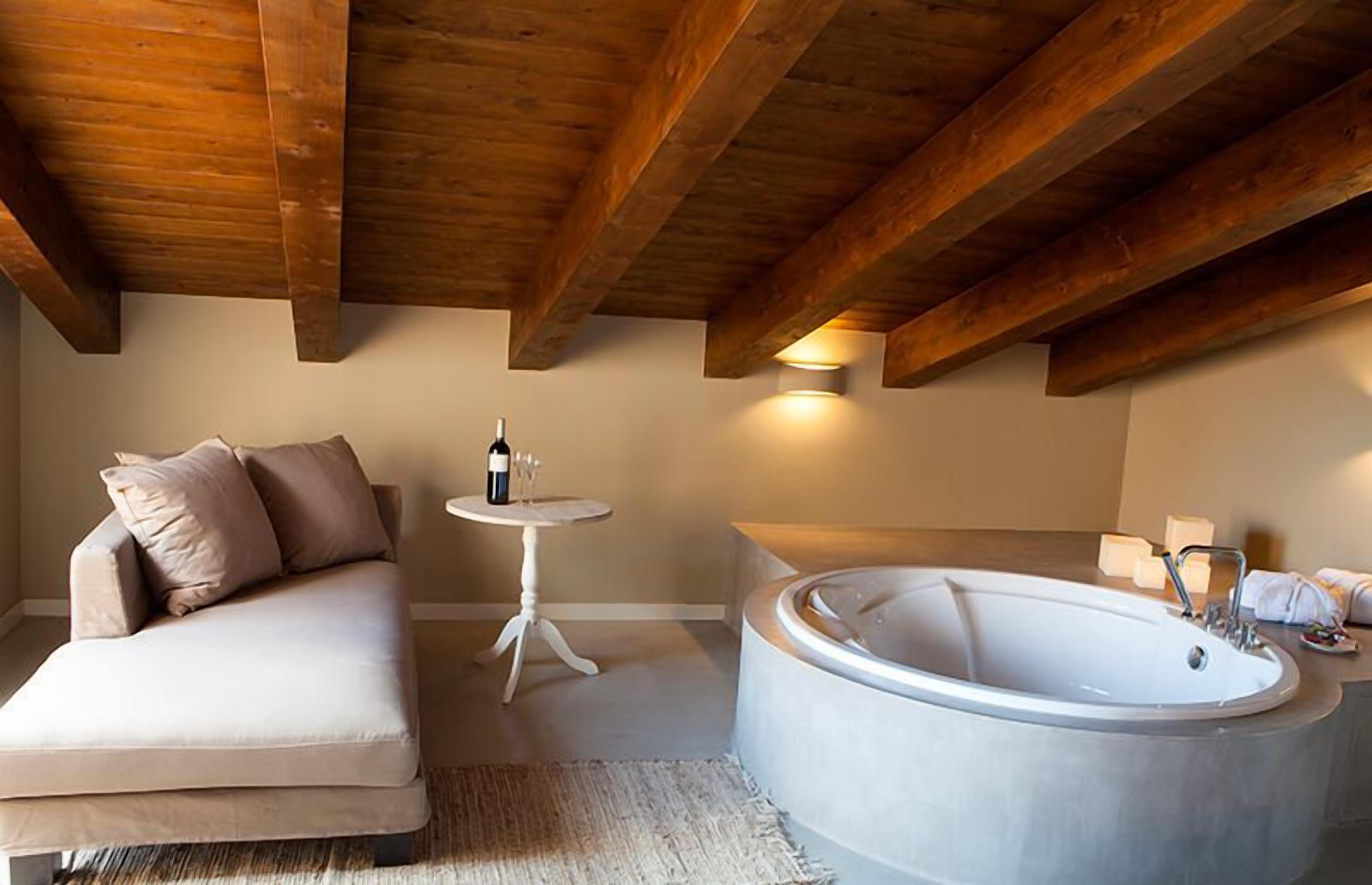 Una habitación acogedora con techo de madera, una bañera grande, un sofá y una pequeña mesa con una botella y vasos.