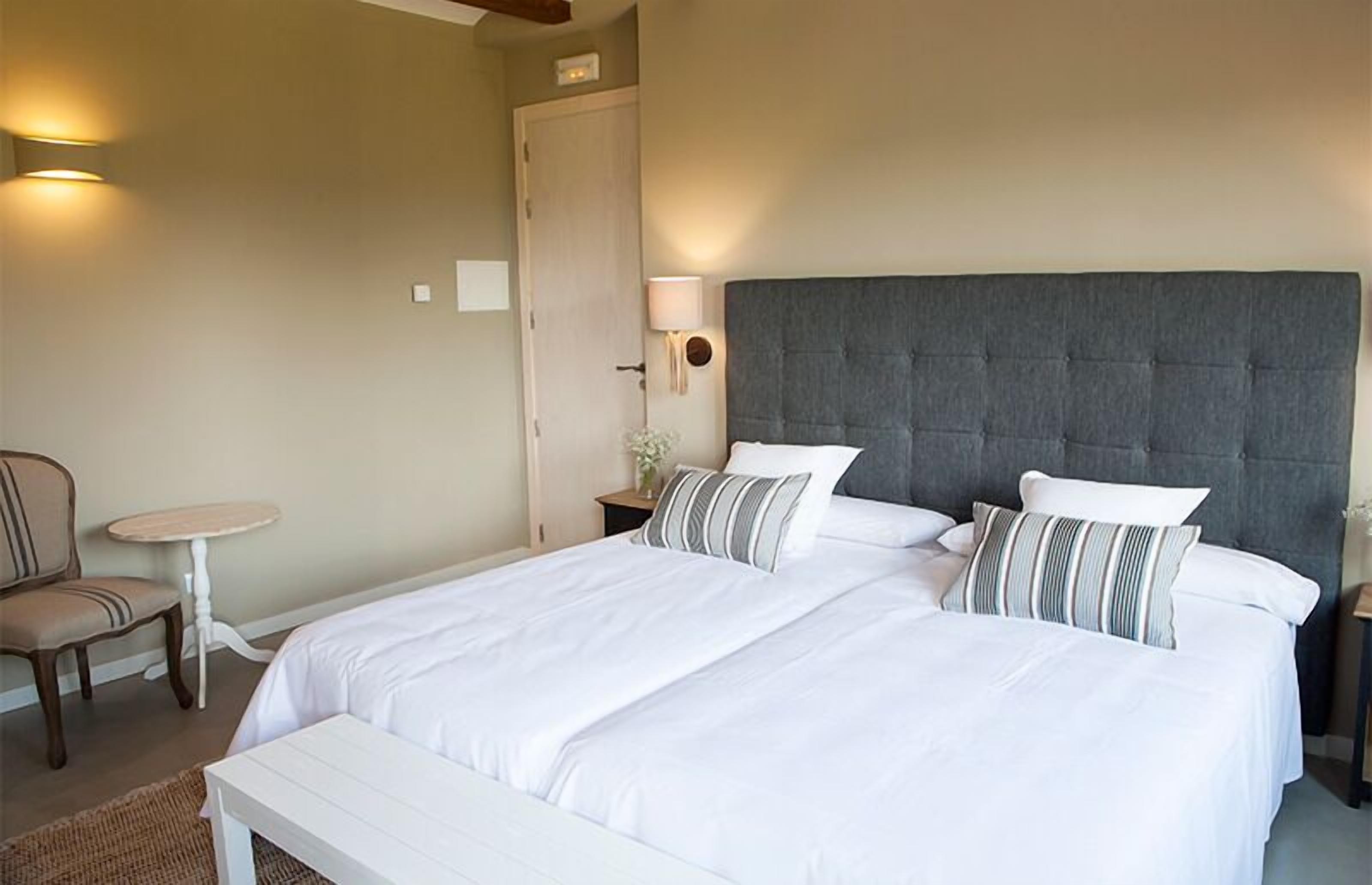 A neatly made bed with white linens, a grey upholstered headboard, and striped pillows. There is a small round table with a chair to the left, and a bedside table with a lamp and a vase of flowers.