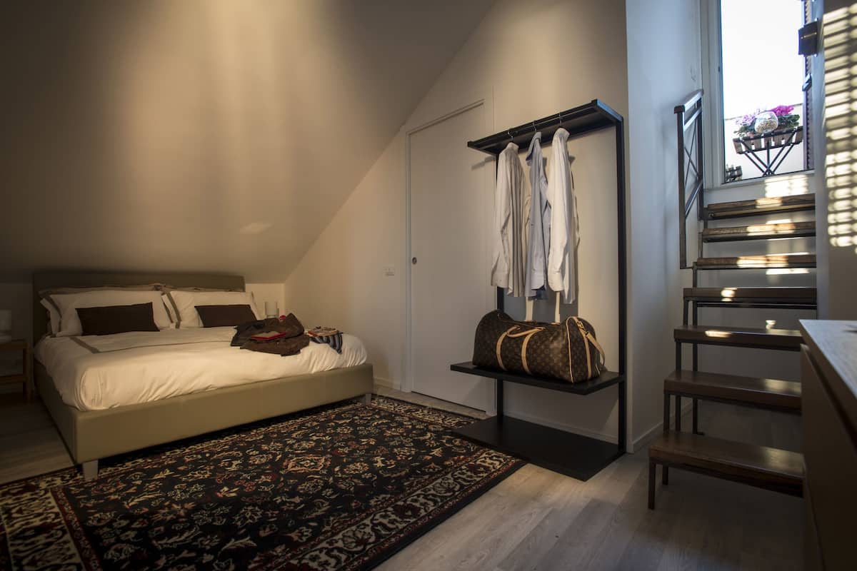 Une chambre avec un lit, un tapis à motifs sombres, une étagère suspendue avec des vêtements et un sac, et un escalier offrant une vue.