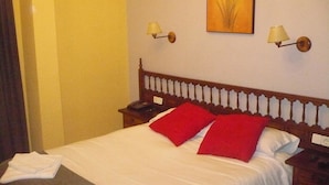A hotel room with a bed, two red pillows, a nightstand with a lamp, a telephone, and a painting on the wall. - Hotel Castillo de Uña (Una)