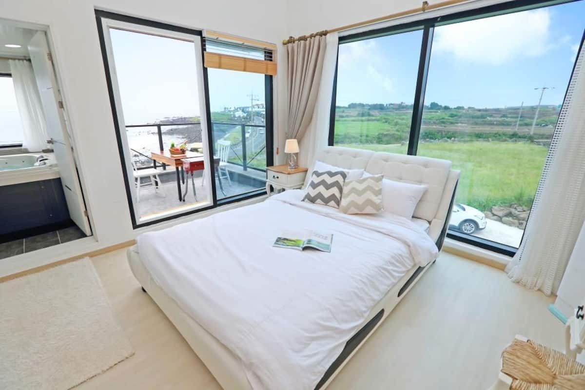 Une chambre à coucher avec un grand lit, une petite table avec une lampe et une vue sur la campagne, visible à travers de grandes fenêtres.
