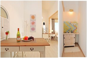 Een eetgedeelte met een houten tafel, een fles wijn en een schaal fruit. Achtergrond toont een kleine witte tafel met een lamp en een stoel. De kamer heeft een witte muur met drie ingelijste foto's.