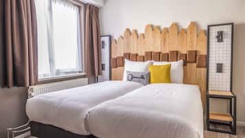 A hotel room with two beds, a wooden headboard, a radiator, a window with curtains, and a small table with a number sign.