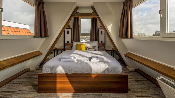 A bedroom with a sloped ceiling, wooden beams, a bed with white bedding, a yellow pillow, and a view of the city through large windows.