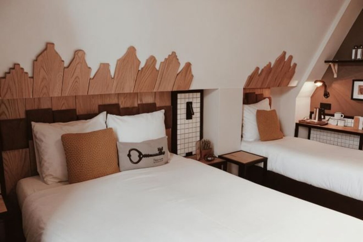 Habitación de hotel con dos camas, cabecero de madera, mesita de noche y una mesita auxiliar.