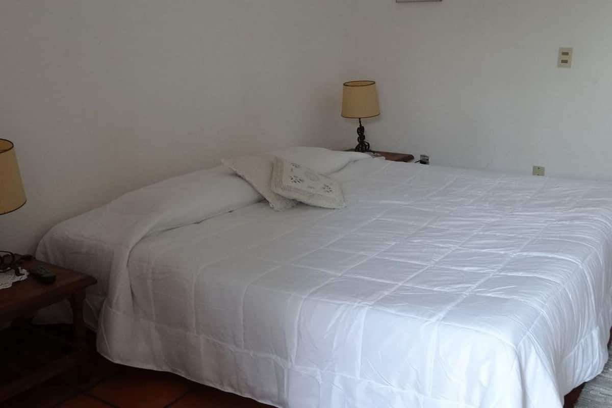 A neatly made bed with a white duvet and a decorative pillow, a bedside table with a lamp, and a wall-mounted light fixture.