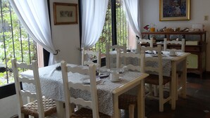 A dining area with white tablecloths, wooden chairs, and a view of greenery outside. - Posada de la Flor (Colonia del Sacramento)