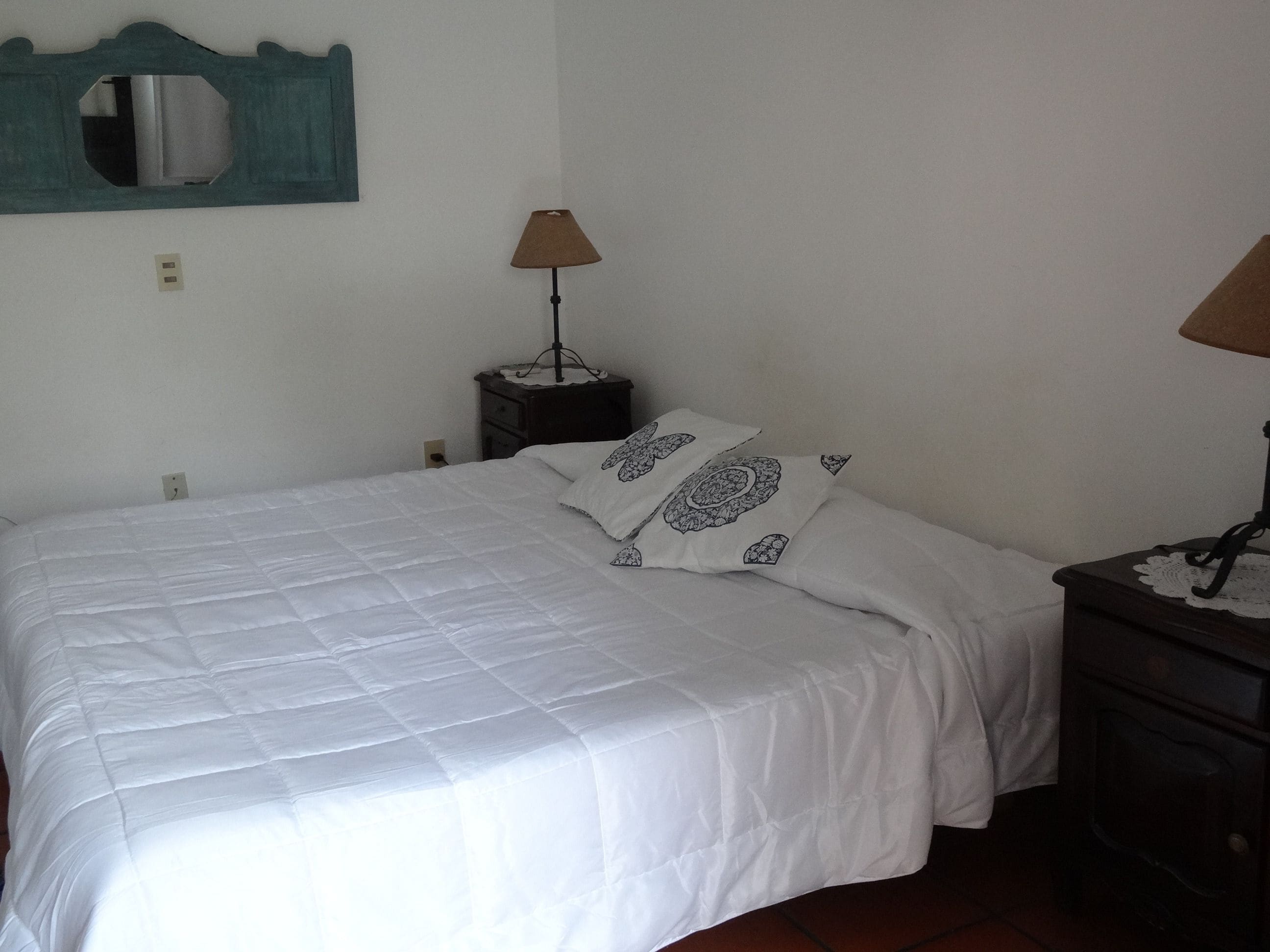 Una cama bien tendida con ropa blanca y almohadas estampadas, una mesita de noche pequeña con una lámpara, y un espejo fijado a la pared.