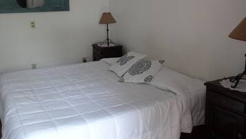 Una cama bien tendida con ropa blanca y almohadas estampadas, una mesita de noche pequeña con una lámpara, y un espejo fijado a la pared.