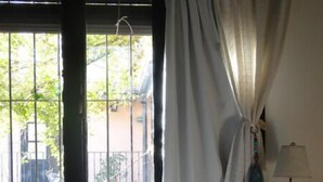 A bedroom with a bed, a nightstand, a lamp, and a window with curtains. - Posada de la Flor (Colonia del Sacramento)