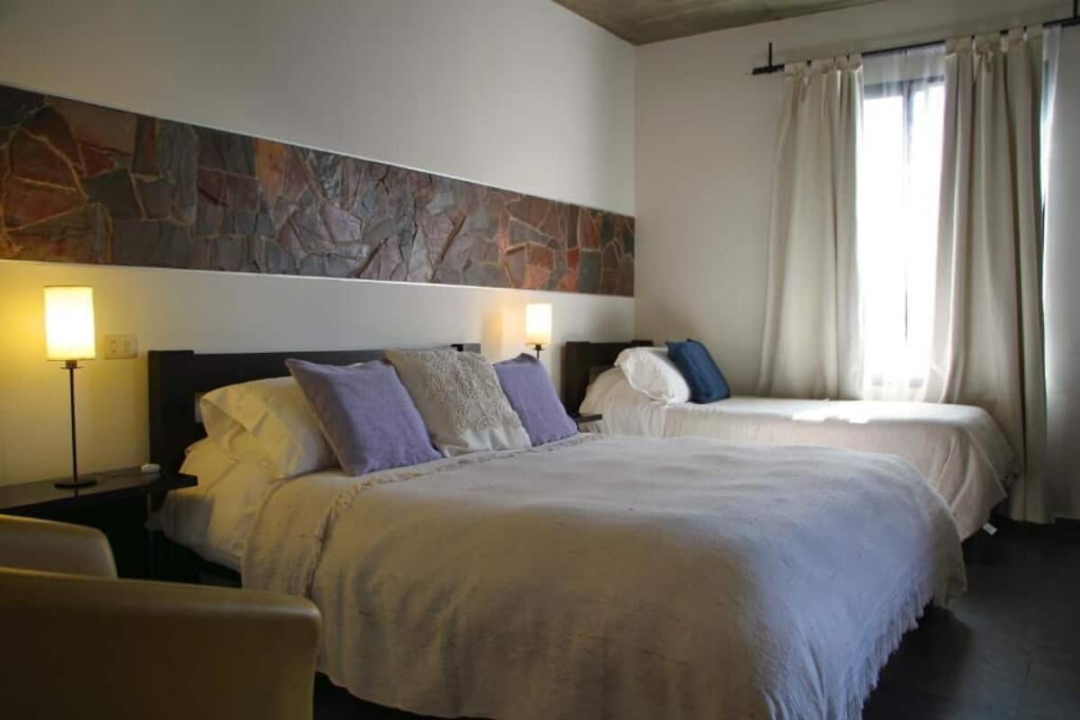 A hotel room with a large bed, a smaller bed, a nightstand with a lamp, a stone wall, and a window with curtains.