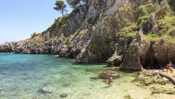A rocky coastline with clear turquoise water, a sandy beach, and a person sitting on a rock. - L'Agrumeto (Diso)