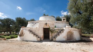 A small, white, dome-shaped building with a stone entrance and steps leading up to it, surrounded by trees and gravel. - Le Site (Ostuni)