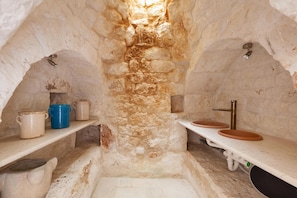 A stone-walled bathroom with two sinks, a shelf with jars, and a small window. - Le Site (Ostuni)