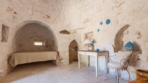 A stone-walled room with a bed, a desk, and a chair. - Le Site (Ostuni)