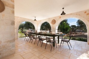 An outdoor dining area with a stone table and chairs, surrounded by arches and a view of the garden. - Le Site (Ostuni)