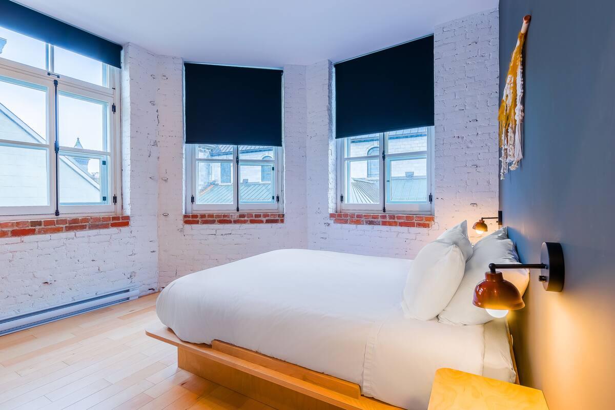 Ein modernes Schlafzimmer mit einem großen Bett, zwei Fenstern mit Jalousien, einer Ziegelwand und einer Wandleuchte.