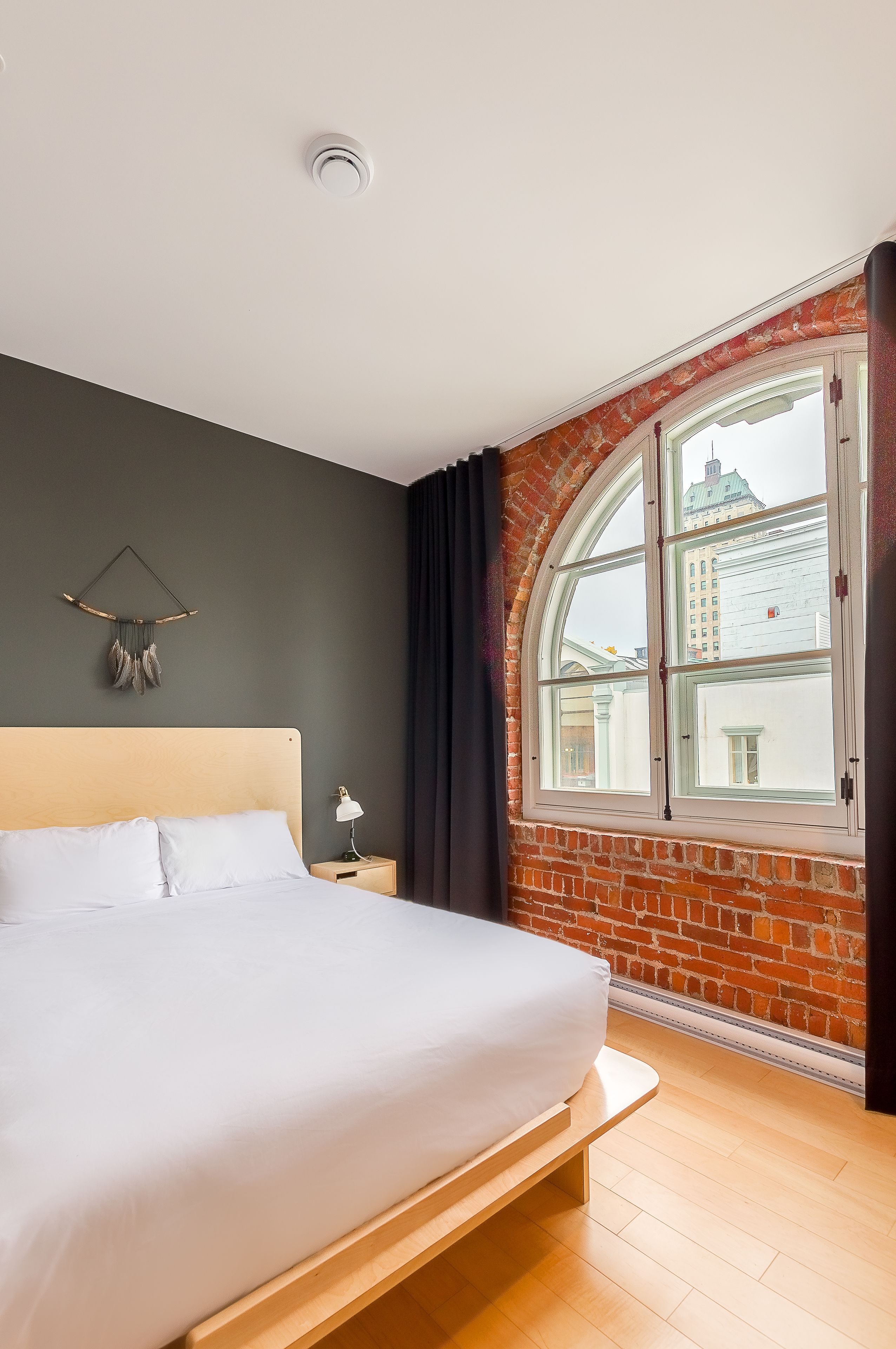 A bedroom with a bed, a nightstand, a lamp, a window with a view of buildings, and a decorative wall hanging.