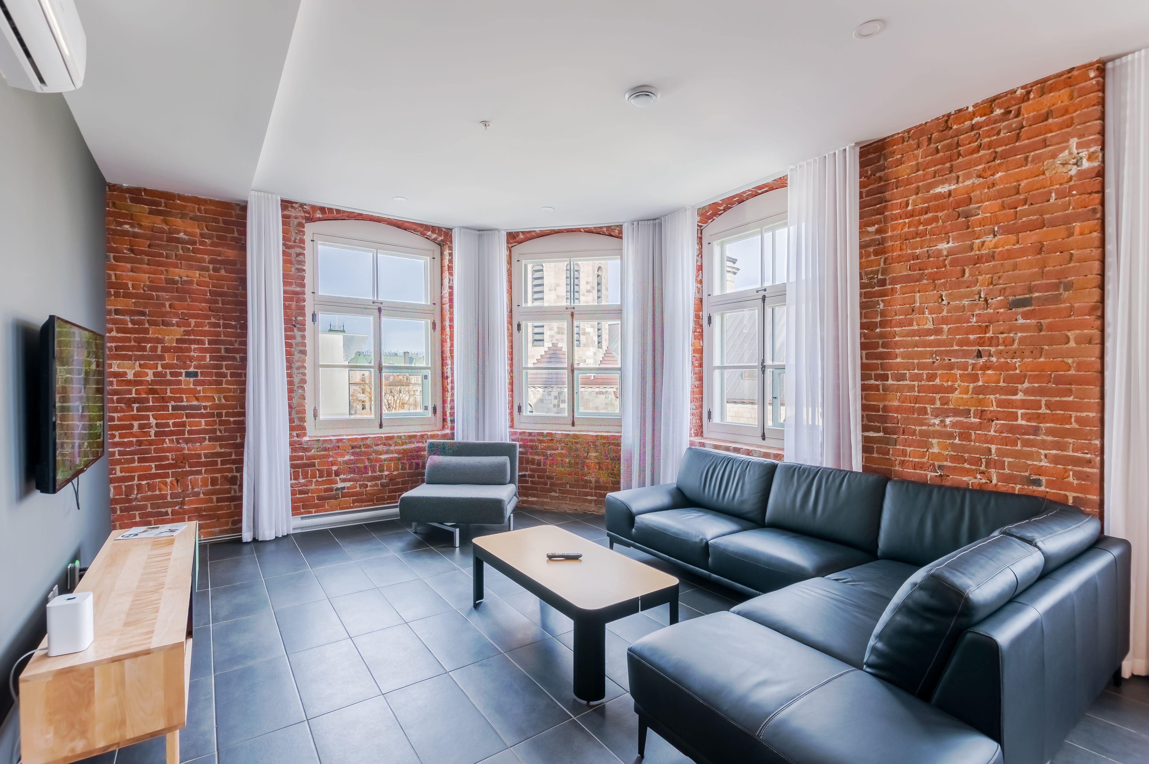 A modern living room with a brick wall, large windows, a black leather sofa, a wooden coffee table, and a grey armchair.