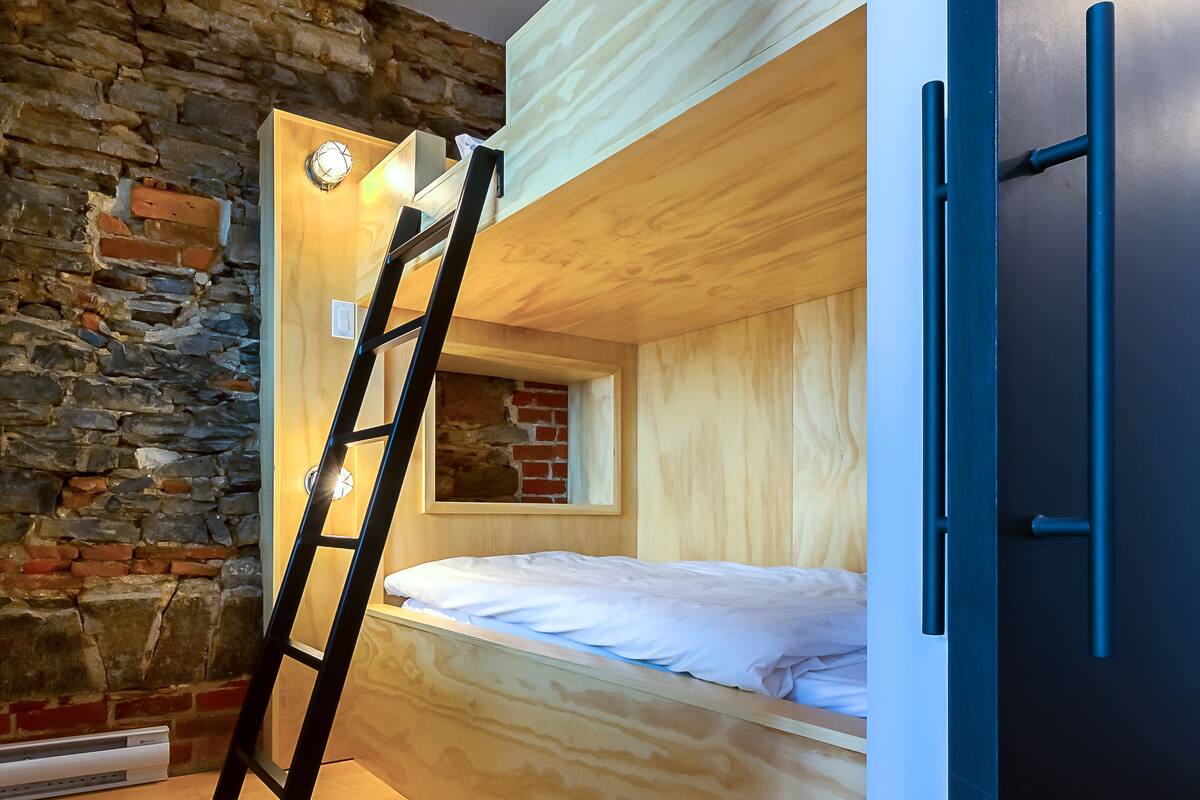 Ein kleines Zimmer mit Etagenbett, einer Leiter, einer Steinsäule und Holzboden.