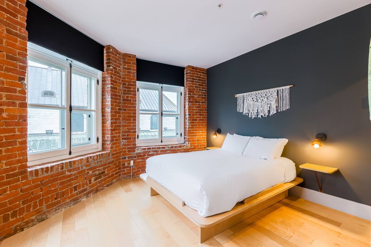 Ein Schlafzimmer mit Backsteinwand, großem Fenster, einem Bett und einer wandhängenden Makramee-Dekoration.