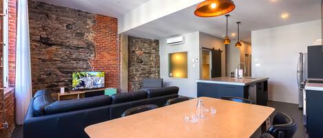 A modern living space with a stone wall, a black leather sofa, a wooden dining table, and a kitchen area.