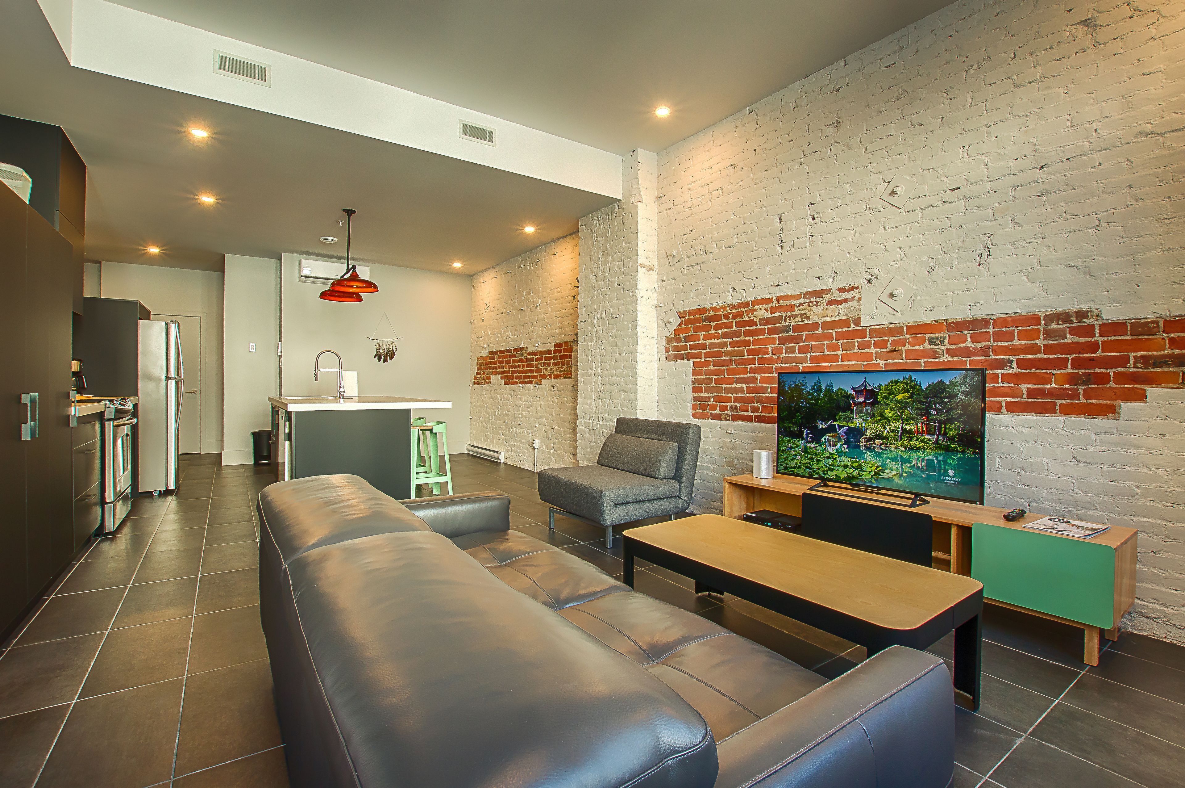 Un salon moderne avec un canapé en cuir, une table basse en bois et un téléviseur fixé à un mur en briques.