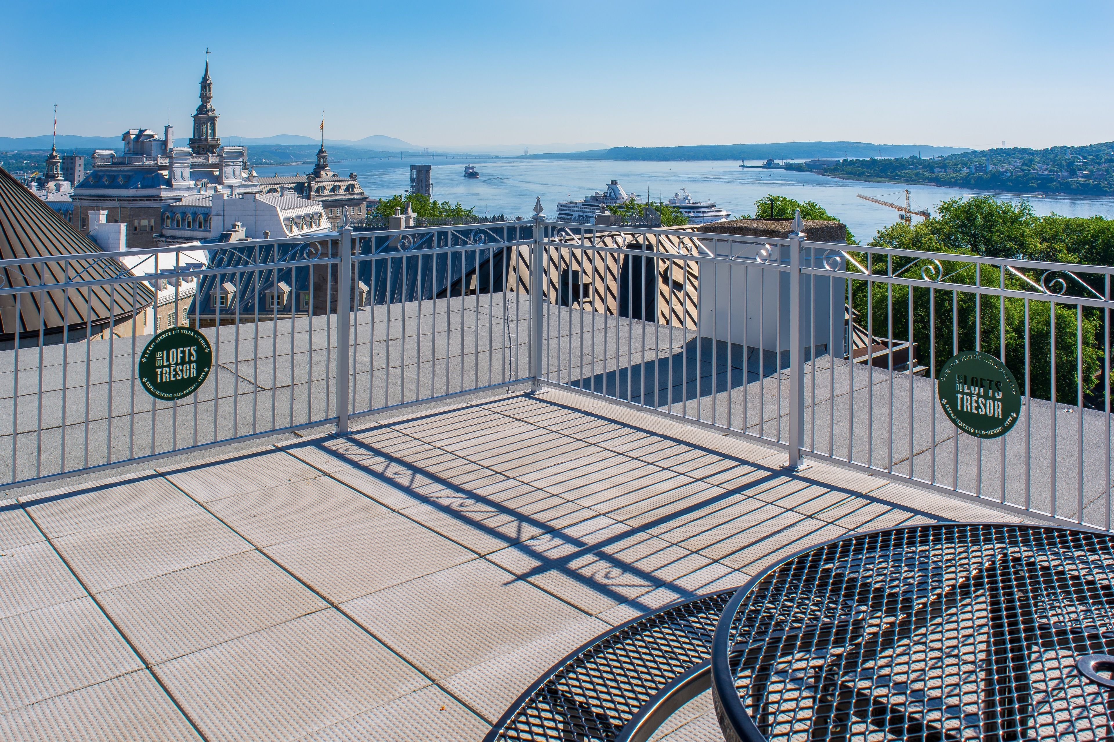 En tagterrasse med gelænder, et rundt bord og udsigt over en flod og bybillede.