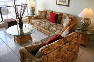 Uma sala de estar com sofá, duas mesas laterais, uma mesa de centro de vidro e um vaso com galhos.