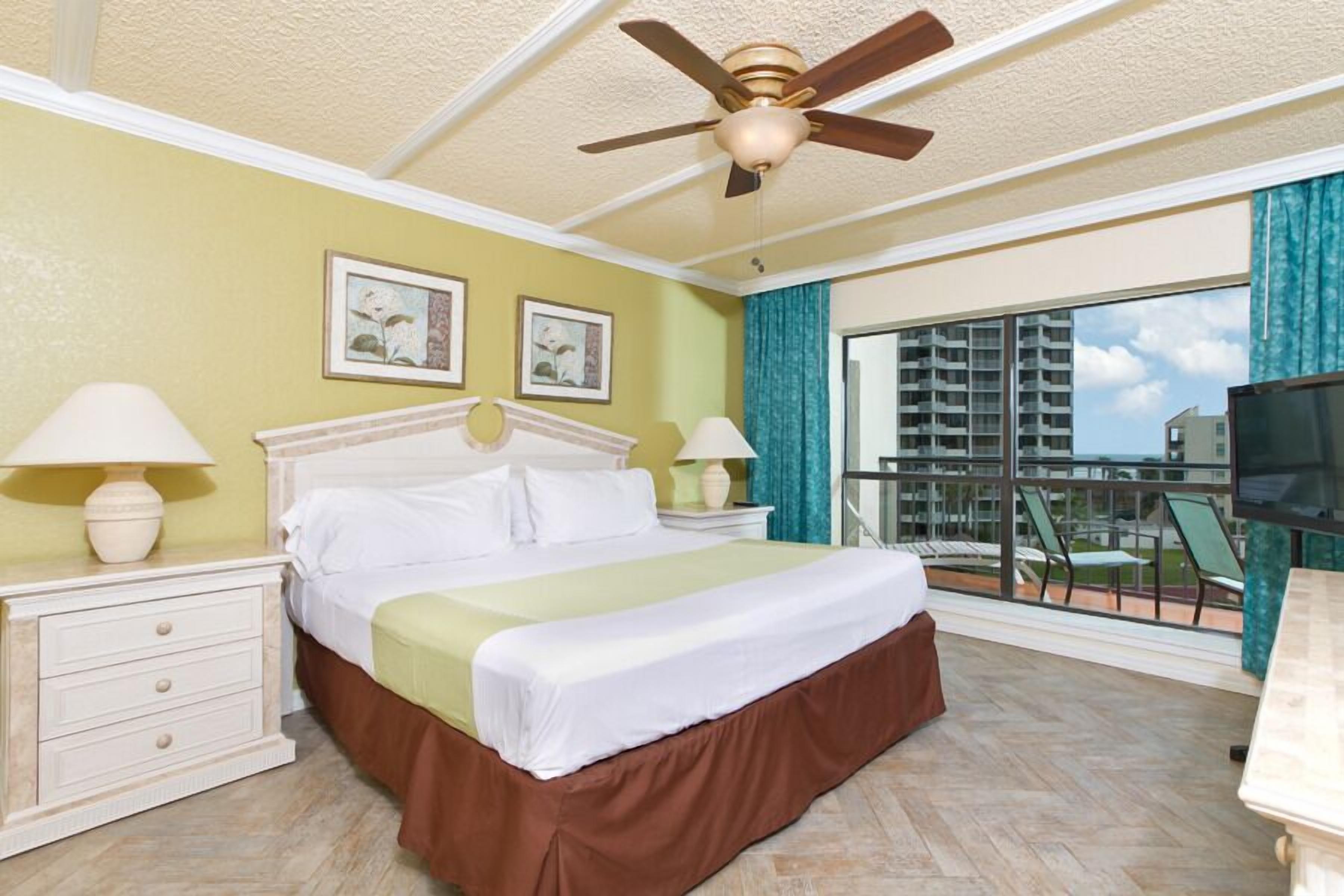 Une chambre d’hôtel comprenant un lit, deux tables de chevet avec lampes, un ventilateur de plafond, un balcon avec des chaises et un téléviseur.